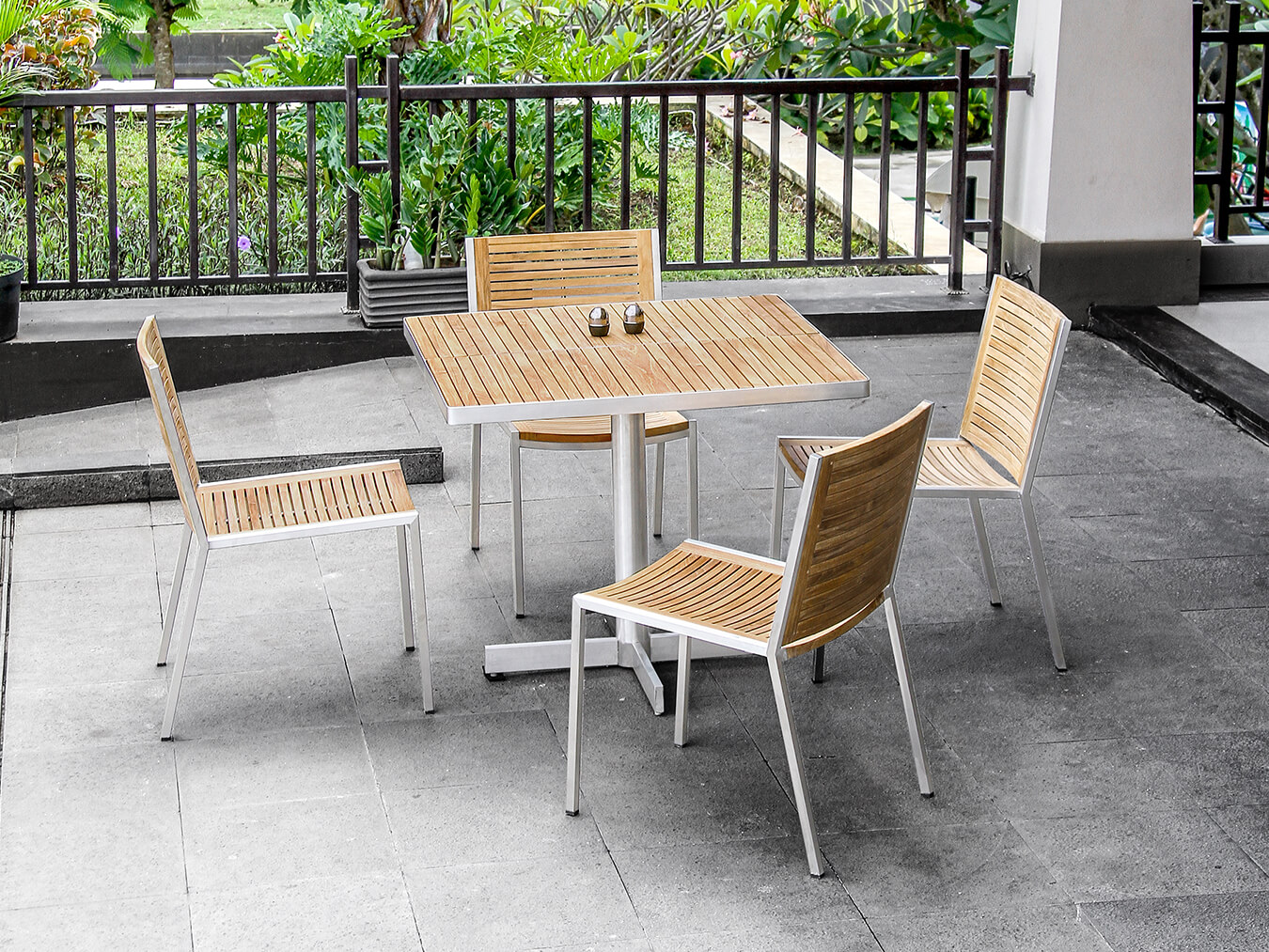 Loom Crafts Outdoor Garden Furniture An outdoor patio set featuring a square wooden table and four matching wooden chairs with metal frames, situated on a tiled surface near a garden area.