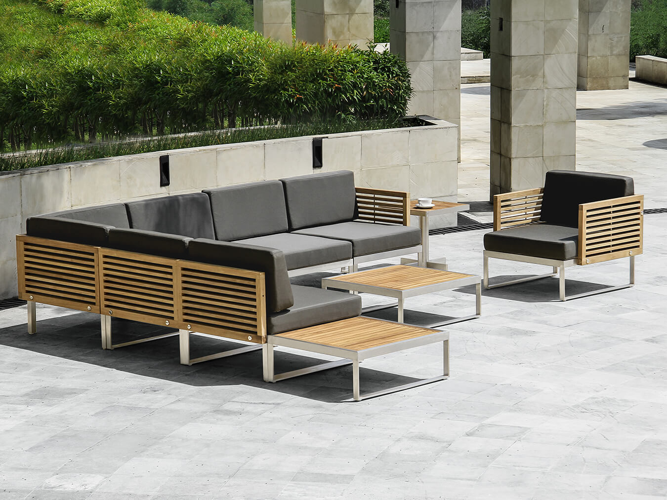 Loom Crafts Outdoor Garden Furniture Outdoor seating area with modern wooden and metal furniture, including a sectional sofa, a single armchair, and two small coffee tables, set on a grey tiled patio surrounded by greenery.